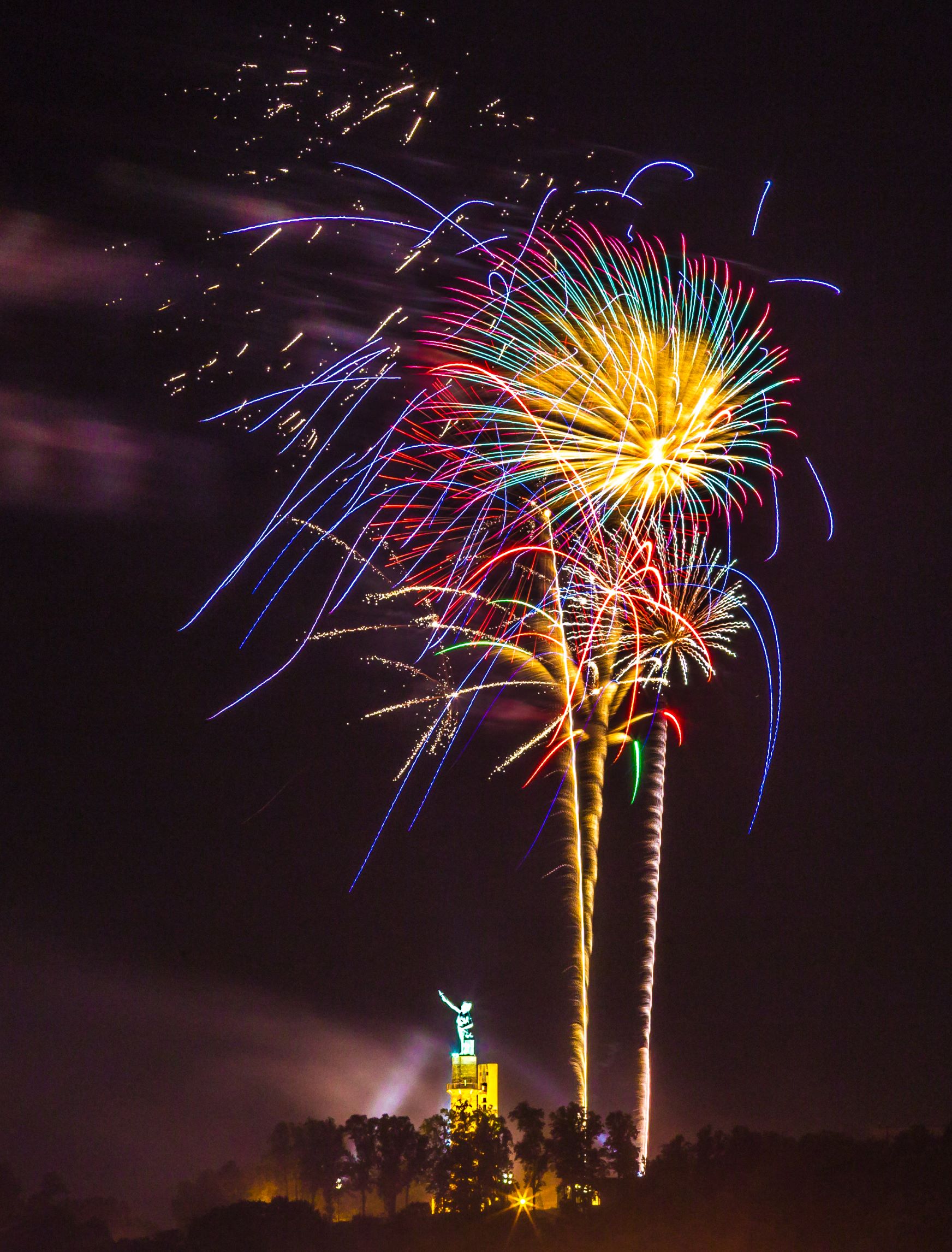 Pink, green, yellow, and blue fireworks over the statue of Vulcan with trees in the foreground