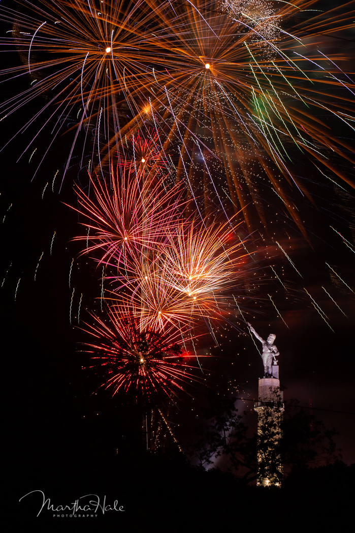 Red, pink, and orange fireworks over the statue of Vulcan with trees in the foreground. 