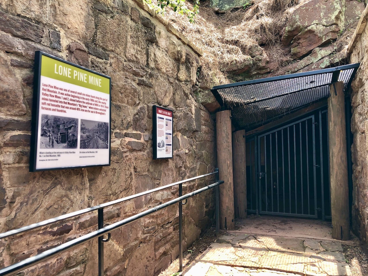 Walkway leading up to mine entrance showing left hand side of rock walls, now with signage with info about mine. Right portion shows black iron gate installed at entrance to mine. 