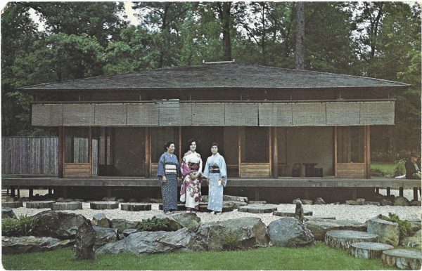 Three women dressed in traditional Japanese Kimonos, stand in front of a brown wooden structure. They are surrounded by large decorative rocks