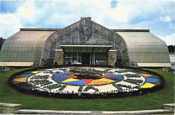 Large, circular flower garden in the shape of a clock in front of a glass building, probably a greenhouse